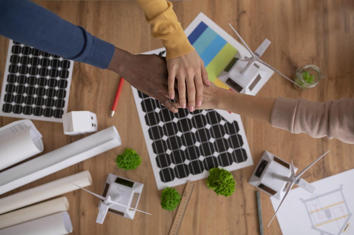 Trois personnes se tiennent la main au dessus d'une table sur laquelle se trouvent des schémas de panneaux solaires, des maquettes d'éoliennes et des plans d'architecture, suggérant un travail d'équipe dans le cadre d'un projet d'énergie renouvelable.