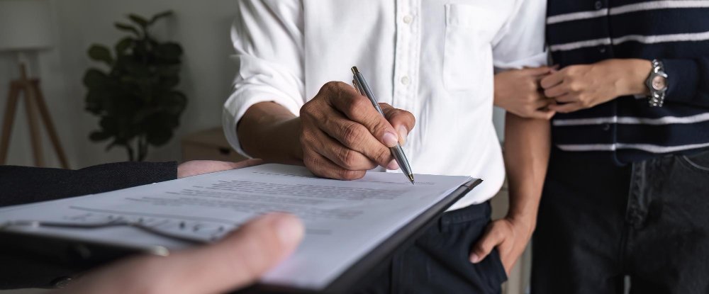 Une personne signe un document sur un bloc-notes tandis qu'une autre se tient à proximité en lui tenant le bras.