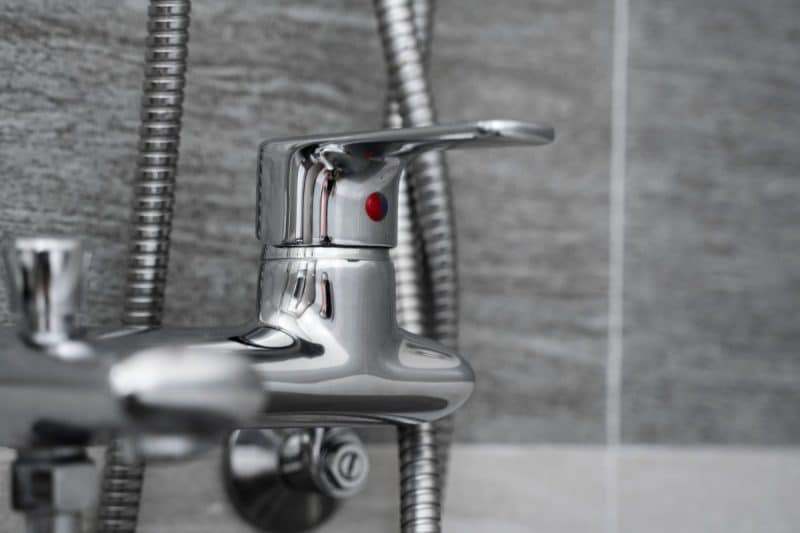 Gros plan d'un robinet de salle de bains chromé brillant avec poignée à levier, installé devant un mur carrelé gris. Le robinet est relié à un tuyau flexible.
