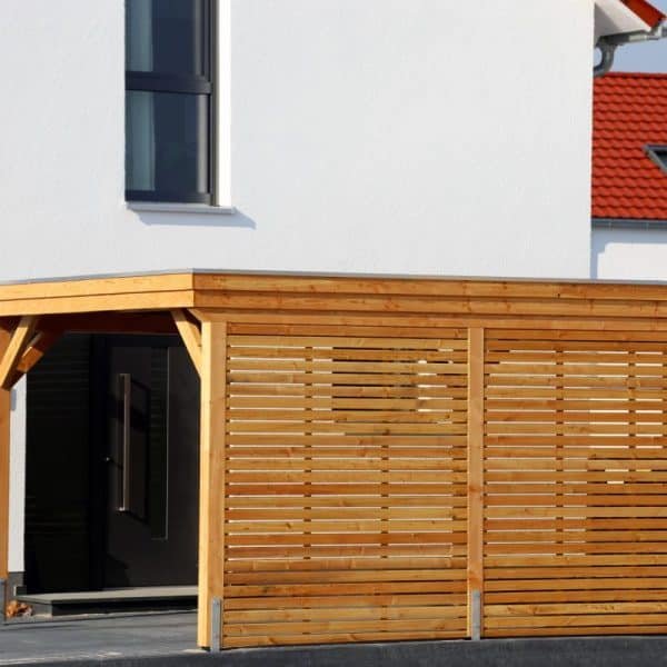 Abri d'extérieur avec ossature en bois et murs à lattes attaché à une maison blanche moderne avec de grandes fenêtres et un toit rouge.