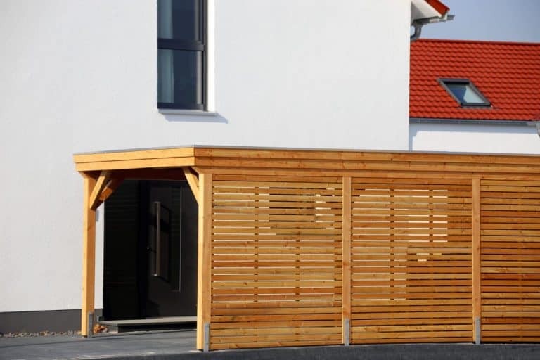 Abri d'extérieur avec ossature en bois et murs à lattes attaché à une maison blanche moderne avec de grandes fenêtres et un toit rouge.