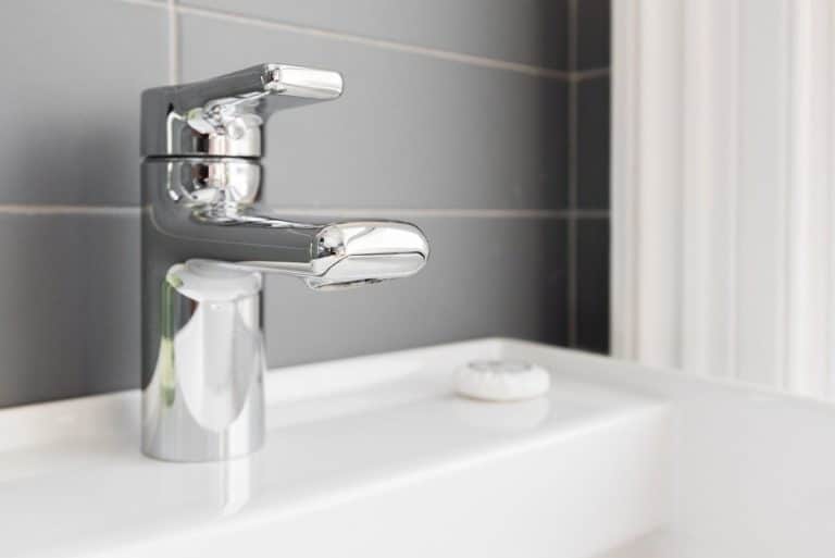 Un robinet chromé brillant monté sur un évier blanc avec un dosseret carrelé gris et un objet rond blanc à proximité.