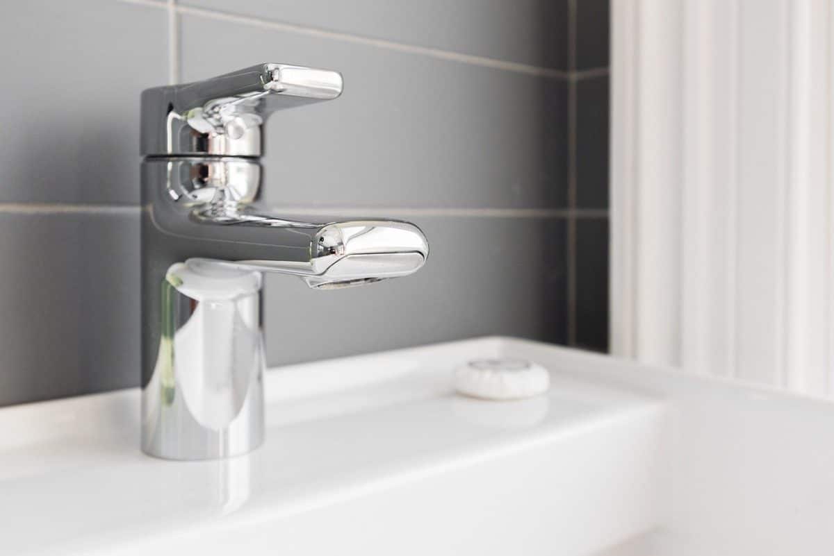Un robinet chromé brillant monté sur un évier blanc avec un dosseret carrelé gris et un objet rond blanc à proximité.