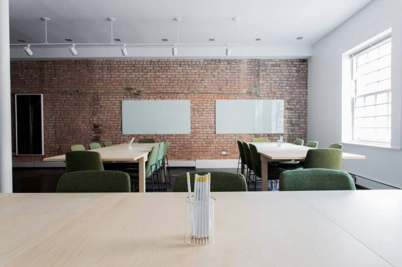 Une salle de conférence moderne avec des tables en bois, des chaises vertes et un mur en briques. Deux tableaux blancs sont fixés au mur. Un verre avec des crayons est posé sur la table au premier plan.
