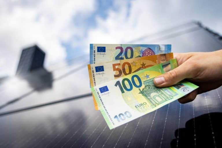 Une main tenant des billets en euros devant un panneau solaire avec un fond de ciel bleu.