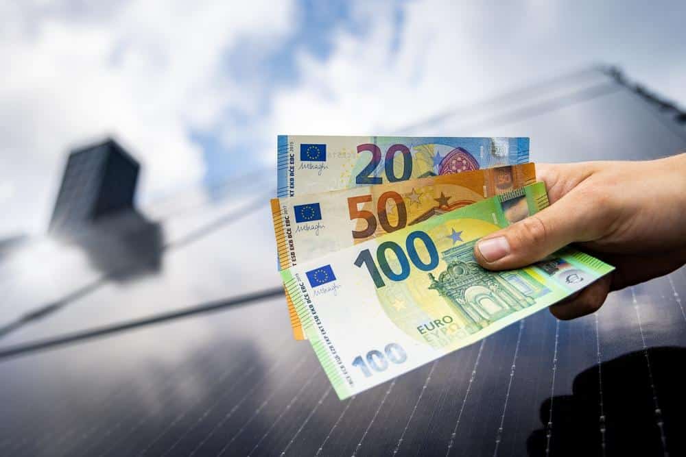 Une main tenant des billets en euros devant un panneau solaire avec un fond de ciel bleu.