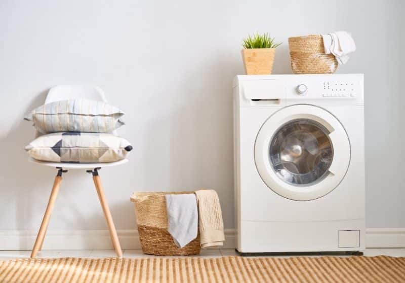 Une machine à laver à côté d'une chaise avec des coussins, un panier à linge avec des vêtements et un panier tressé avec des plantes et des serviettes au dessus de la machine dans une buanderie bien rangée.