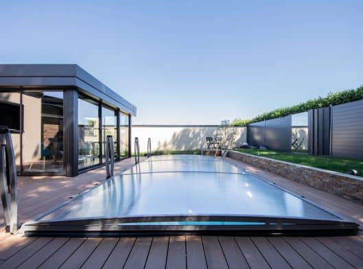 Une piscine extérieure moderne avec une couverture transparente et rétractable se trouve à côté d'un bâtiment aux parois de verre et entourée d'une cour clôturée avec de l'herbe.