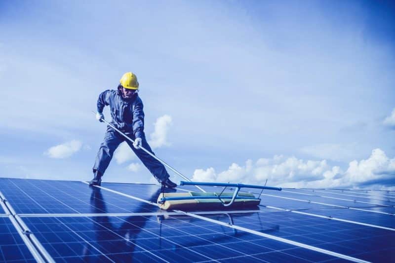 Un travailleur en équipement de protection nettoie les panneaux solaires sur un toit sous un ciel bleu avec des nuages épars.