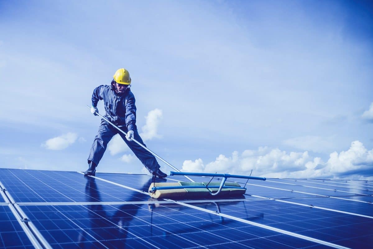 Un travailleur en équipement de protection nettoie les panneaux solaires sur un toit sous un ciel bleu avec des nuages épars.