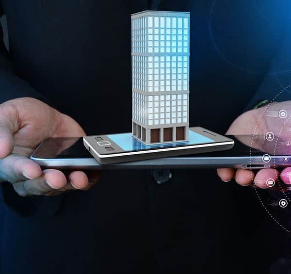 Une personne tient une tablette et un smartphone avec un modèle numérique d'un bâtiment affiché au dessus, symbolisant la technologie dans l'immobilier ou la gestion intelligente des bâtiments.