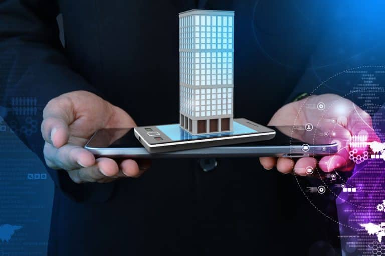 Une personne tient une tablette et un smartphone avec un modèle numérique d'un bâtiment affiché au dessus, symbolisant la technologie dans l'immobilier ou la gestion intelligente des bâtiments.