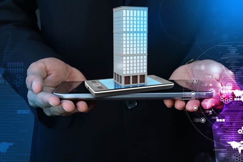 Une personne tient une tablette et un smartphone avec un modèle numérique d'un bâtiment affiché au dessus, symbolisant la technologie dans l'immobilier ou la gestion intelligente des bâtiments.