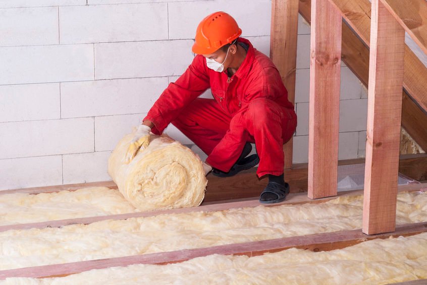 Un ouvrier en uniforme rouge et casque orange installe une isolation en fibre de verre sur un plancher entre des poutres en bois à l'intérieur d'un bâtiment en construction.