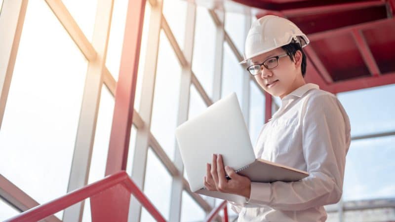 Jeune ingénieur asiatique avec un casque utilisant un ordinateur portable dans un environnement industriel avec de grandes fenêtres.