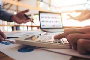 Gros plan de mains utilisant une calculatrice et discutant de documents sur une table, avec un ordinateur portable en arrière plan.