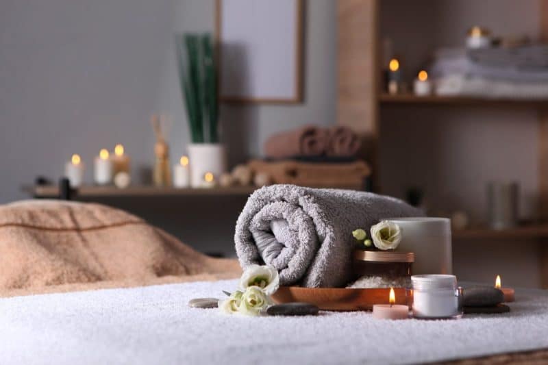 Une serviette grise soigneusement roulée avec des fleurs blanches repose sur un lit de spa, entourée de bougies allumées, de lotions et de pierres dans une pièce faiblement éclairée avec des serviettes pliées et une plante en arrière plan.