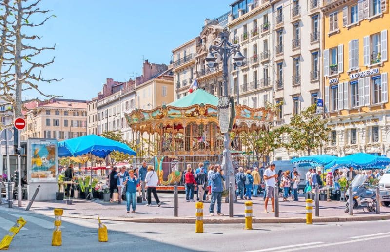 Une rue animée avec des gens rassemblés autour d'un carrousel orné entouré de bâtiments colorés et d'auvents bleus par une journée ensoleillée.
