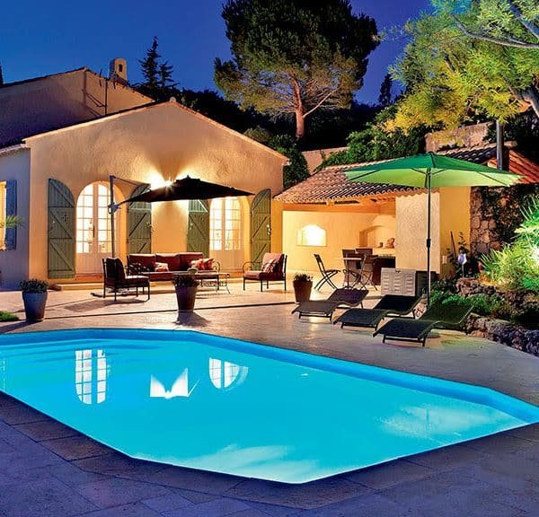 Une maison avec une piscine éclairée au crépuscule, entourée d'arbres et de mobilier extérieur sous des parasols.