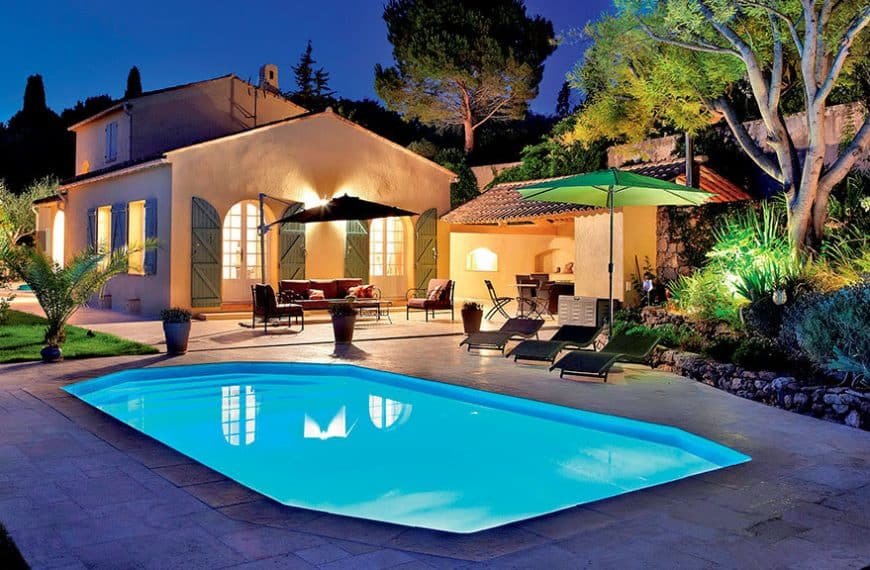 Une maison avec une piscine éclairée au crépuscule, entourée d'arbres et de mobilier extérieur sous des parasols.