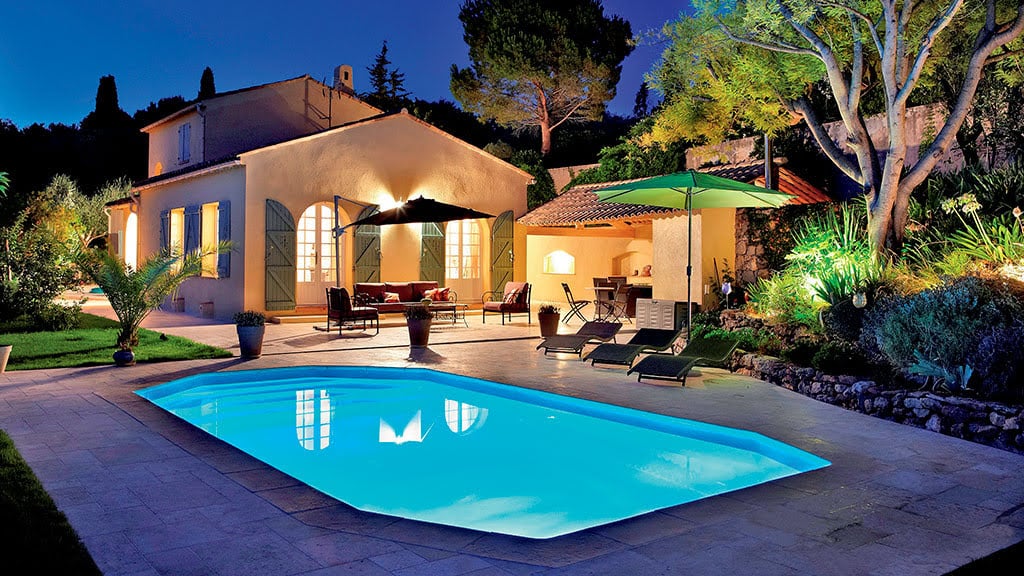 Une maison avec une piscine éclairée au crépuscule, entourée d'arbres et de mobilier extérieur sous des parasols.