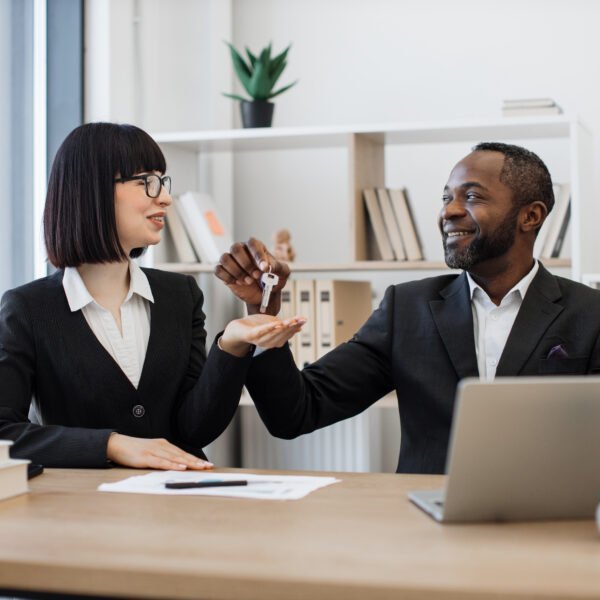Deux professionnels en tenue de ville sont assis à un bureau ; l'un remet un trousseau de clés à l'autre, tandis que des documents et un ordinateur portable sont visibles sur la table.