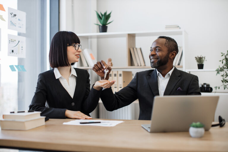 Deux professionnels en tenue de ville sont assis à un bureau ; l'un remet un trousseau de clés à l'autre, tandis que des documents et un ordinateur portable sont visibles sur la table.