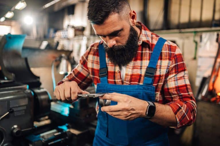Un homme en chemise à carreaux et salopette bleue travaille avec un pied à coulisse dans un atelier, mesurant une pièce métallique. Des machines et des outils sont visibles en arrière plan.