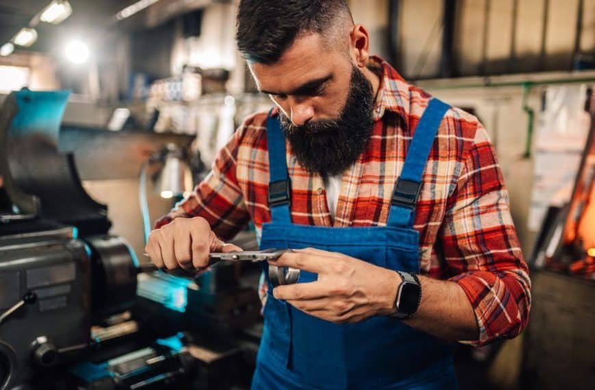Un homme en chemise à carreaux et salopette bleue travaille avec un pied à coulisse dans un atelier, mesurant une pièce métallique. Des machines et des outils sont visibles en arrière plan.