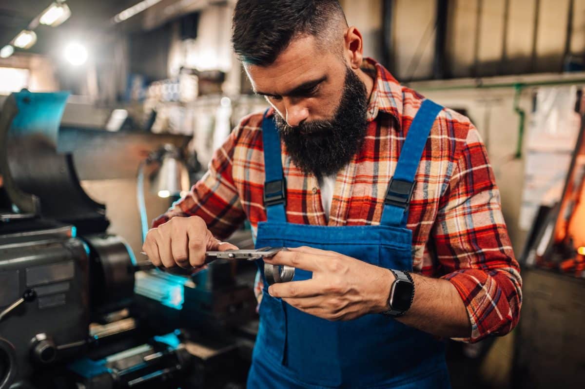 Un homme en chemise à carreaux et salopette bleue travaille avec un pied à coulisse dans un atelier, mesurant une pièce métallique. Des machines et des outils sont visibles en arrière plan.