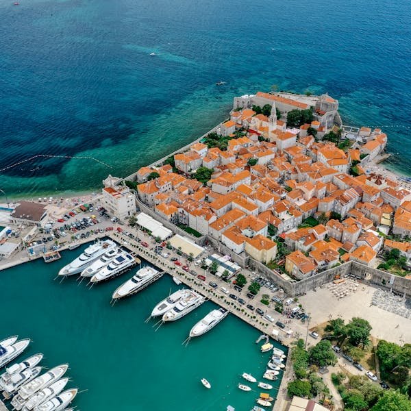 Vue aérienne d'une ville côtière avec des bâtiments aux toits orangés, un port de plaisance rempli de yachts et une eau bleue limpide.