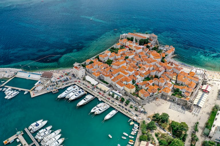 Vue aérienne d'une ville côtière avec des bâtiments aux toits orangés, un port de plaisance rempli de yachts et une eau bleue limpide.