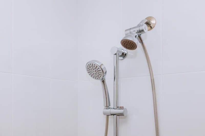 Une configuration de douche avec une pomme de douche montée, une pomme de douche à main sur un rail réglable et des tuyaux flexibles contre un mur carrelé blanc.