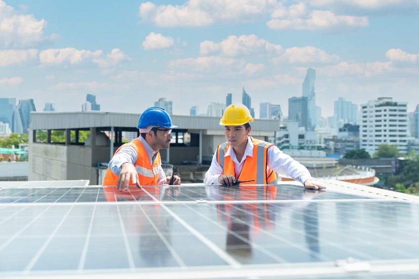 Deux ouvriers portant des gilets de sécurité et des casques inspectent des panneaux solaires sur un toit avec un horizon de la ville en arrière plan.