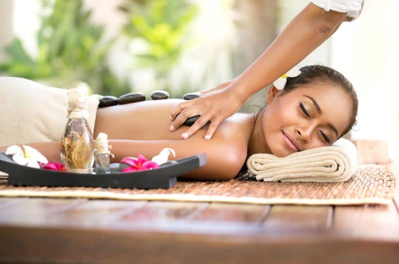 Une personne recevant un massage aux pierres chaudes sur le dos, allongée sur une table de massage avec un oreiller en serviette. Un plateau avec des bouteilles d'huile et des fleurs est placé à côté d'eux.
