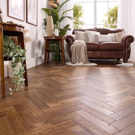 Un salon avec un parquet à chevrons, un canapé en cuir marron, des tables d'appoint ornées de plantes et de grandes fenêtres laissant entrer la lumière naturelle.