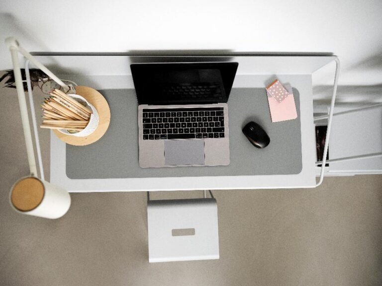 Vue d'ensemble d'un bureau minimaliste avec un ordinateur portable, une lampe, un pot de crayons, une souris et deux petits blocs-notes sur un tapis de bureau gris.