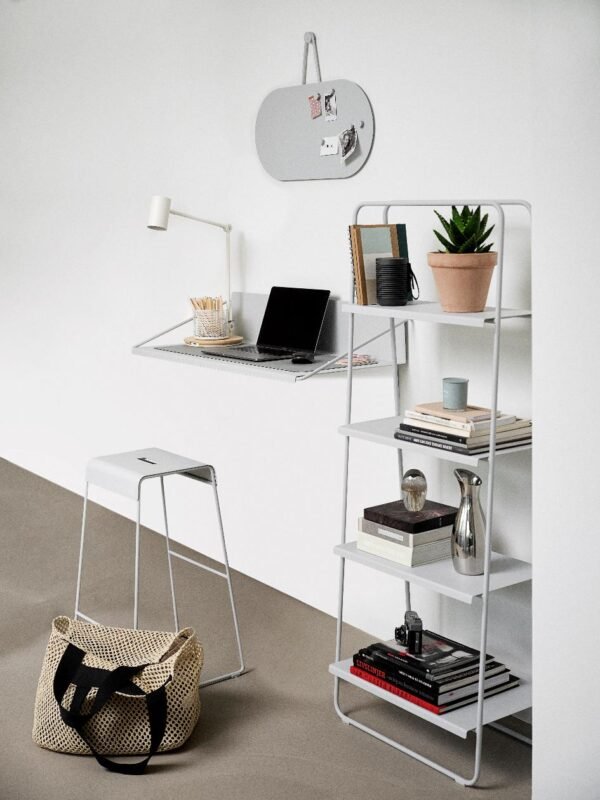 Bureau à domicile minimaliste composé d'un bureau mural, d'un ordinateur portable, d'un tabouret, d'une étagère contenant des livres et des objets de décoration, d'une plante en pot et d'un grand sac tissé posé sur le sol.