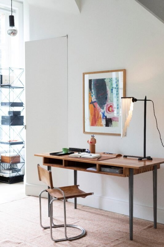 Un bureau en bois surmonté d'une lampe, de fournitures artistiques et d'une œuvre d'art abstraite encadrée est placé à côté d'une chaise moderne dans une pièce lumineuse et minimaliste dotée d'une fenêtre et d'une étagère.