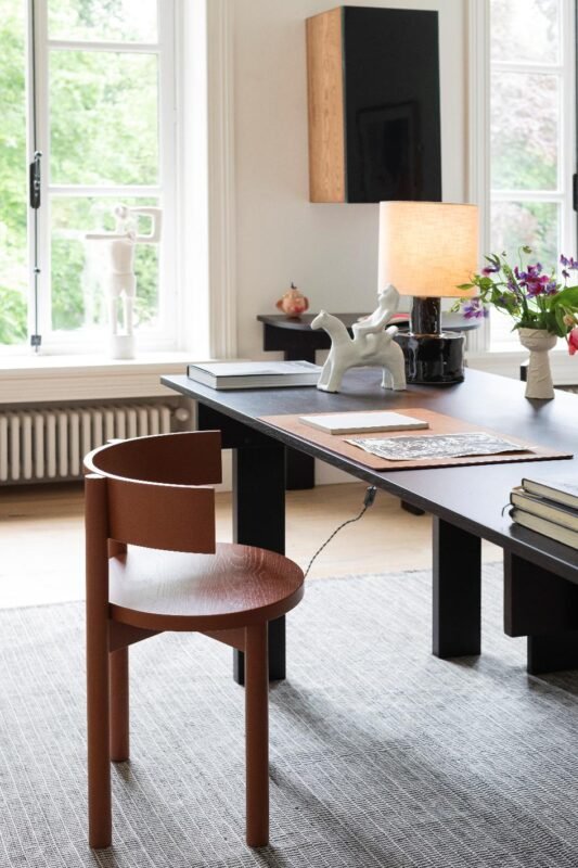 Un bureau moderne avec une chaise en bois incurvée, un bureau sombre, une lampe de table, des livres, un vase avec des fleurs et des sculptures près de grandes fenêtres.