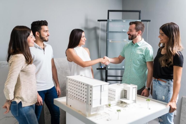 Cinq personnes se tiennent autour d'une table avec une maquette d'architecture ; deux d'entre elles se serrent la main, tandis que les autres observent et sourient dans un bureau moderne.