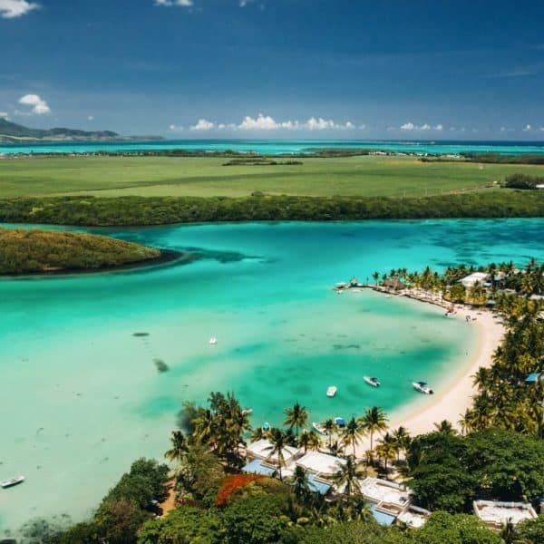 Vue aérienne d'une plage tropicale aux eaux turquoise Ile Maurice, au rivage sablonneux et aux bateaux dispersés. Des palmiers et des paysages verdoyants entourent la zone sous un ciel bleu clair.