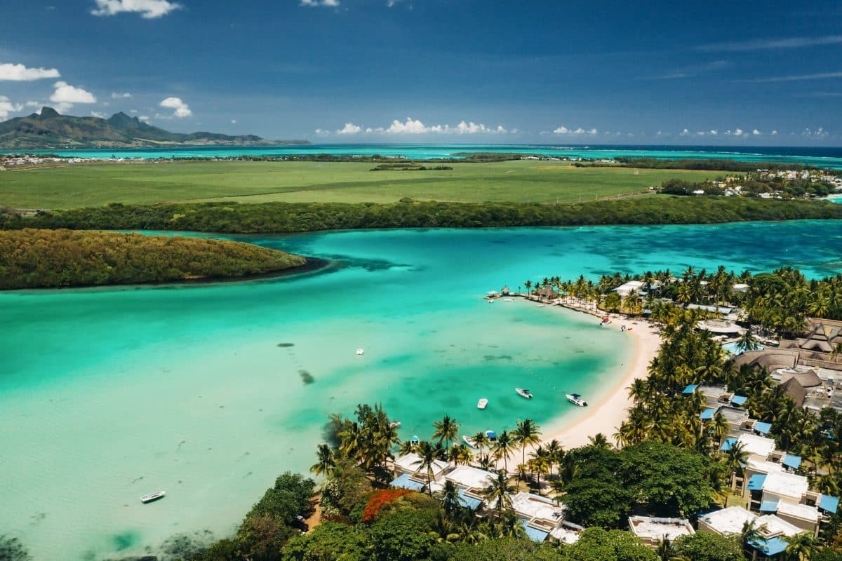 Vue aérienne d'une plage tropicale aux eaux turquoise Ile Maurice, au rivage sablonneux et aux bateaux dispersés. Des palmiers et des paysages verdoyants entourent la zone sous un ciel bleu clair.