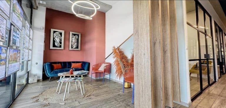 Un hall d'entrée moderne avec un canapé bleu, une table basse ronde et des œuvres d'art sur un mur couleur corail. Il y a des chaises orange et des plantes décoratives près d'un escalier avec des pièces aux parois vitrées sur la droite.