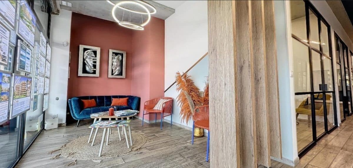 Un hall d'entrée moderne avec un canapé bleu, une table basse ronde et des œuvres d'art sur un mur couleur corail. Il y a des chaises orange et des plantes décoratives près d'un escalier avec des pièces aux parois vitrées sur la droite.