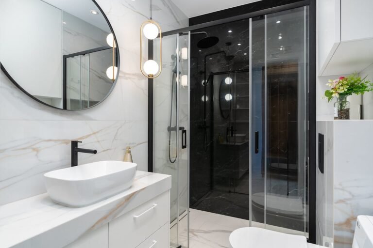 Salle de bains moderne avec un miroir rond, un lavabo, des accessoires noirs, des surfaces en marbre blanc, une cabine de douche en verre et un petit vase de fleurs sur une étagère.
