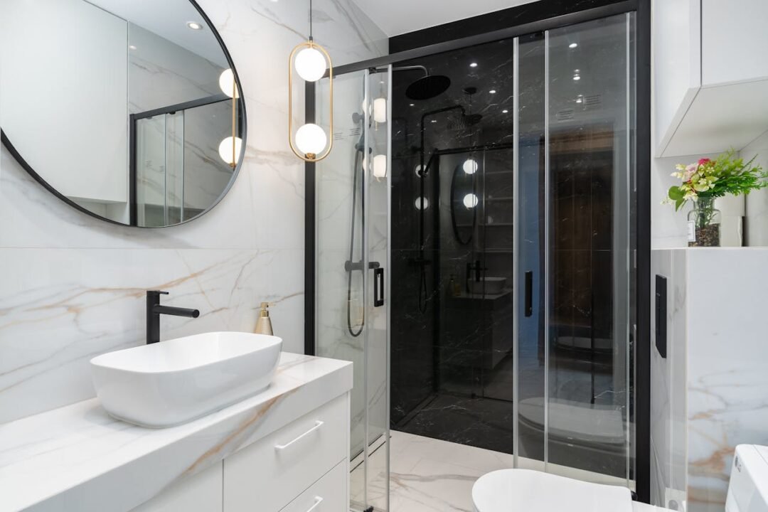 Salle de bains moderne avec un miroir rond, un lavabo, des accessoires noirs, des surfaces en marbre blanc, une cabine de douche en verre et un petit vase de fleurs sur une étagère.