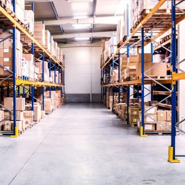 Intérieur de l'entrepôt avec des étagères hautes remplies de diverses boîtes et emballages en carton, et une allée dégagée au centre.