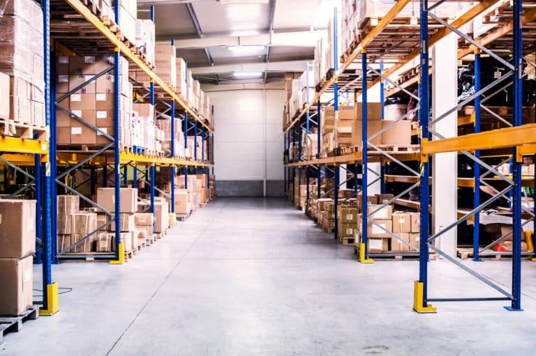 Intérieur de l'entrepôt avec des étagères hautes remplies de diverses boîtes et emballages en carton, et une allée dégagée au centre.