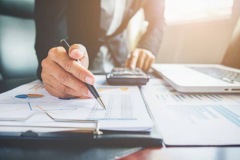 Une personne en tenue de travail examine des documents financiers avec des tableaux et des graphiques, à l'aide d'un stylo et d'une calculatrice, avec un ordinateur portable à proximité sur le bureau.
